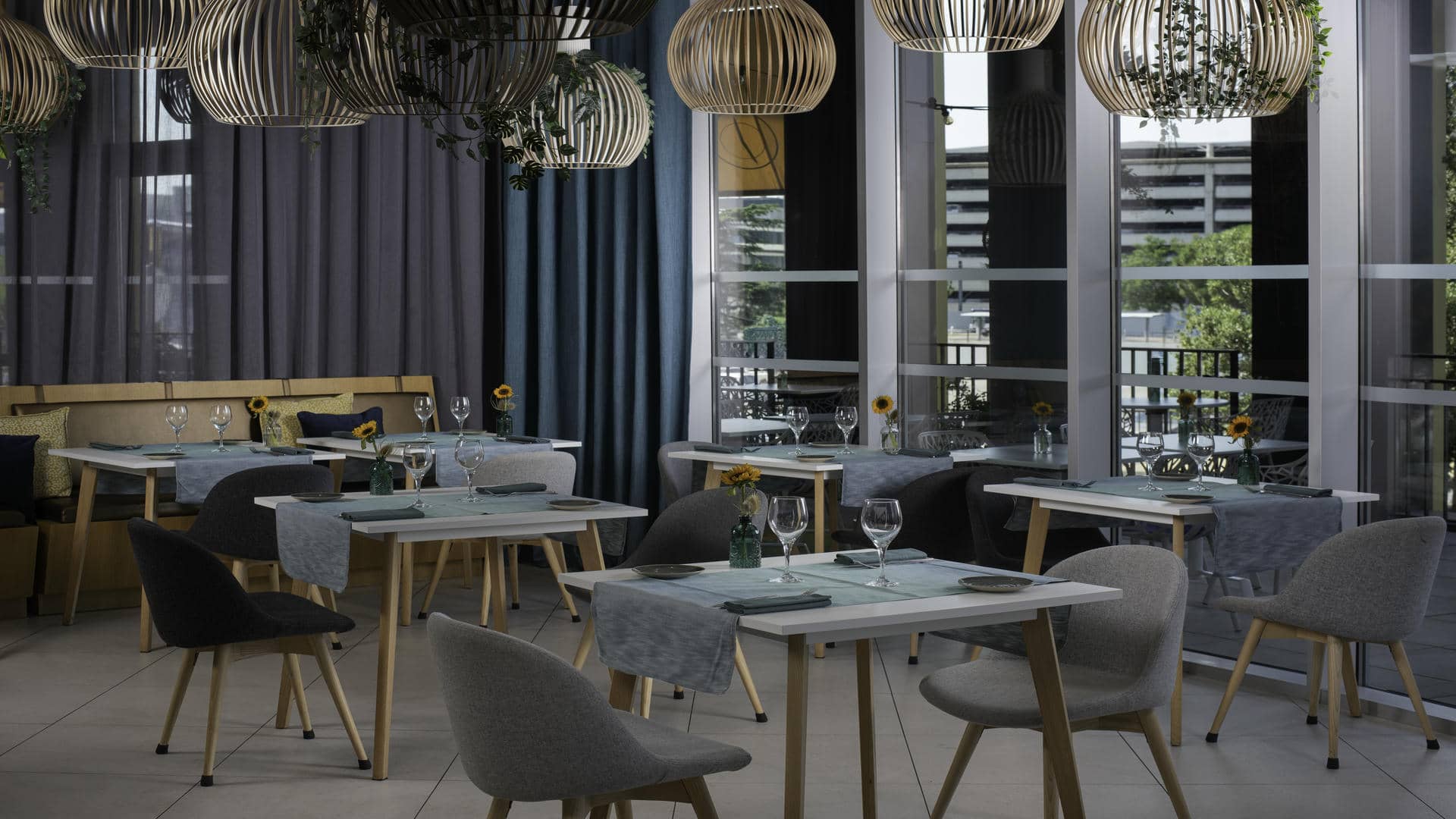 Salle de restaurant aménagée avec plusieurs tables rectangulaires blanches, recouvertes de chemins de table bleus et dressées avec des assiettes, des serviettes en tissu foncé, des verres à pied et de petits vases contenant des tournesols. Les tables sont entourées de fauteuils en tissu gris clair ou gris anthracite, aux pieds en bois clair. Le long du mur gauche, une banquette beige est agrémentée de coussins gris, beiges et bleu foncé. De grandes baies vitrées occupent le côté droit et offrent une vue sur des arbres et des bâtiments. Des rideaux gris foncé et bleu pétrole encadrent les fenêtres. Au plafond, de nombreuses suspensions arrondies en lattes de bois clair et foncé, certaines agrémentées de plantes vertes retombantes, surplombent la salle. Le sol est recouvert de grands carreaux gris clair.
