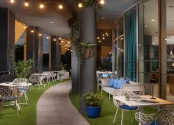 Terrasse de restaurant aménagée avec des tables carrées et des chaises modernes grises aux pieds effet bois gris. Le sol est recouvert de dalles grises, avec des tapis imitation gazon disposés sous certaines tables. Des piliers noirs soutiennent la structure et accueillent des plantes vertes dans des pots en bois. Des guirlandes lumineuses et des luminaires suspendus aux finitions effet bois éclairent l’espace. Certaines tables sont installées le long des baies vitrées du restaurant, tandis que d’autres offrent une vue dégagée sur la ville.