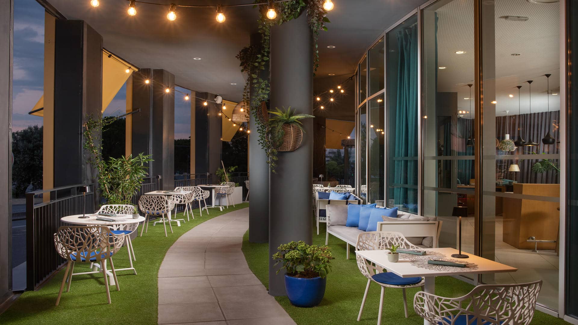 Terrasse de restaurant aménagée avec des tables carrées et des chaises modernes grises aux pieds effet bois gris. Le sol est recouvert de dalles grises, avec des tapis imitation gazon disposés sous certaines tables. Des piliers noirs soutiennent la structure et accueillent des plantes vertes dans des pots en bois. Des guirlandes lumineuses et des luminaires suspendus aux finitions effet bois éclairent l’espace. Certaines tables sont installées le long des baies vitrées du restaurant, tandis que d’autres offrent une vue dégagée sur la ville.