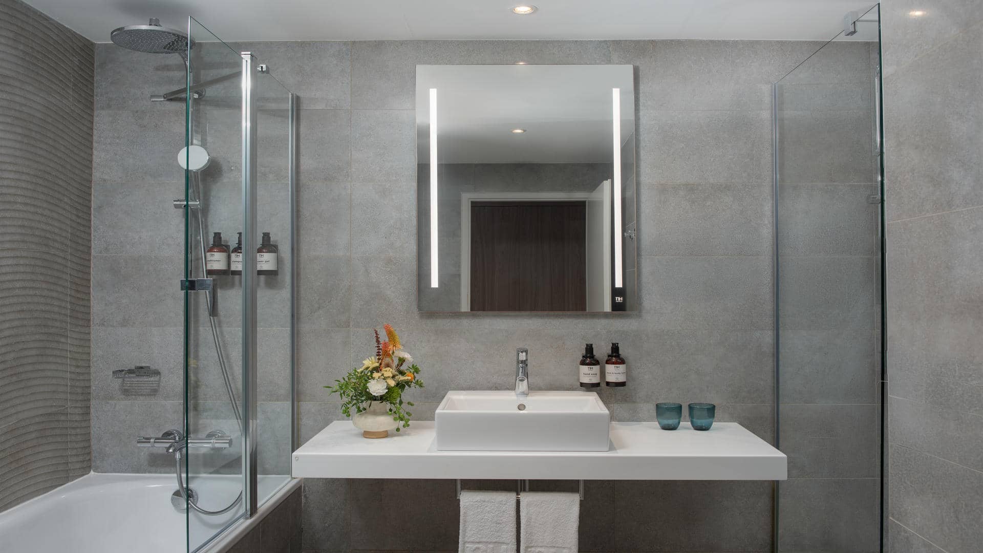 Salle de bain aux murs et au sol recouverts de carrelage gris effet pierre. Une vasque rectangulaire blanche repose sur un meuble en marbre blanc. Au-dessus, un grand miroir rectangulaire lumineux occupe le mur. Un petit vase contenant une composition florale colorée est disposé sur le comptoir. À gauche, une baignoire équipée d’une douche est installée, tandis qu’une douche à l’italienne se trouve sur le côté droit. Des spots lumineux intégrés au plafond éclairent l’ensemble de la pièce.