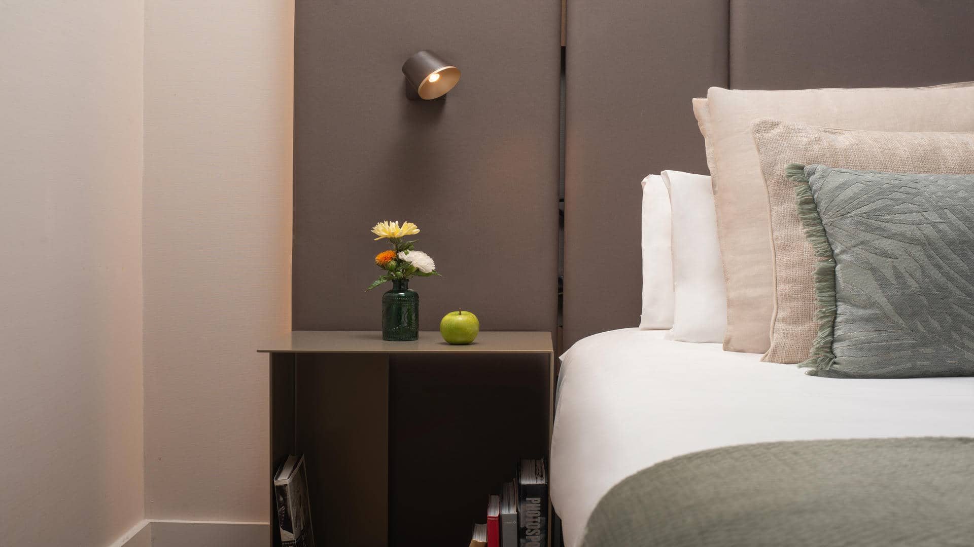 Chambre d’hôtel avec un lit habillé de linge blanc et une tête de lit rembourrée en tissu taupe foncé. Plusieurs oreillers blancs, beiges et gris vert sont disposés contre la tête de lit, assortis à un plaid gris vert posé au pied du lit. À gauche, une table de chevet brun foncé avec une niche contenant des livres accueille un petit vase vert avec des fleurs jaunes, orange et blanches ainsi qu’une pomme verte. Une applique murale noire à lumière chaude est fixée au-dessus de la table. Les murs sont beige clair, en contraste avec la tête de lit foncée.