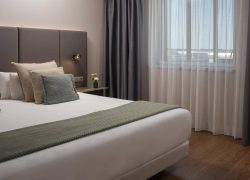 Chambre d’hôtel avec un grand lit double habillé de linge blanc, accompagné de plusieurs oreillers blancs, beiges et gris vert. Une tête de lit rembourrée, composée de panneaux verticaux taupe foncé, occupe le mur derrière le lit. Un plaid texturé vert sauge est disposé en travers du lit. À droite, une petite table de chevet foncée accueille un livre et un vase vert contenant quelques fleurs jaunes et blanches. Une applique murale noire à lumière chaude est fixée au-dessus. Une large fenêtre est habillée de voilages blancs et de rideaux occultants gris anthracite. Le sol est recouvert d’un parquet brun et les murs sont beige clair.