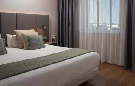 Chambre d’hôtel avec un grand lit double habillé de linge blanc, accompagné de plusieurs oreillers blancs, beiges et gris vert. Une tête de lit rembourrée, composée de panneaux verticaux taupe foncé, occupe le mur derrière le lit. Un plaid texturé vert sauge est disposé en travers du lit. À droite, une petite table de chevet foncée accueille un livre et un vase vert contenant quelques fleurs jaunes et blanches. Une applique murale noire à lumière chaude est fixée au-dessus. Une large fenêtre est habillée de voilages blancs et de rideaux occultants gris anthracite. Le sol est recouvert d’un parquet brun et les murs sont beige clair.