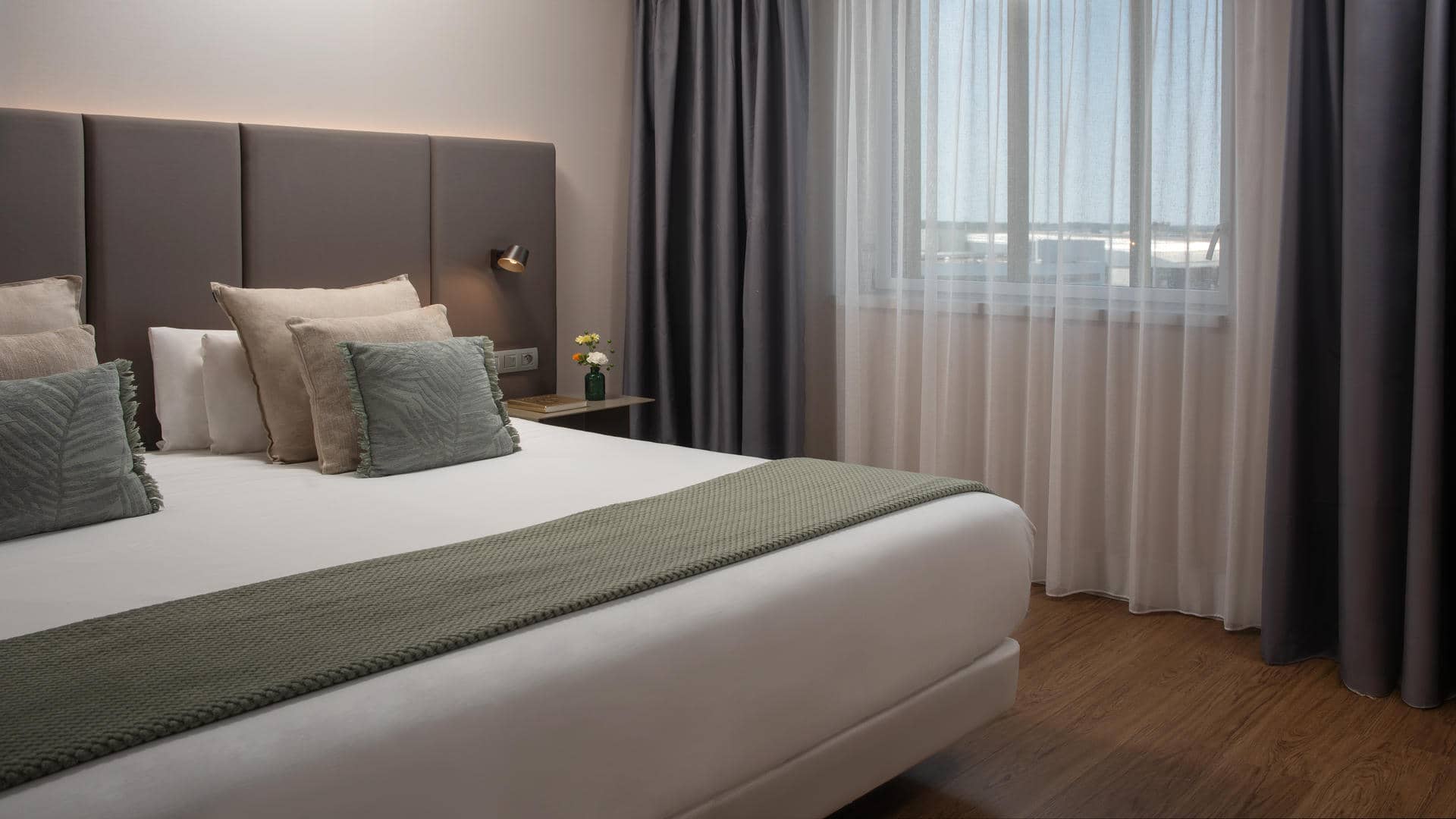 Chambre d’hôtel avec un grand lit double habillé de linge blanc, accompagné de plusieurs oreillers blancs, beiges et gris vert. Une tête de lit rembourrée, composée de panneaux verticaux taupe foncé, occupe le mur derrière le lit. Un plaid texturé vert sauge est disposé en travers du lit. À droite, une petite table de chevet foncée accueille un livre et un vase vert contenant quelques fleurs jaunes et blanches. Une applique murale noire à lumière chaude est fixée au-dessus. Une large fenêtre est habillée de voilages blancs et de rideaux occultants gris anthracite. Le sol est recouvert d’un parquet brun et les murs sont beige clair.