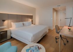 Chambre d’hôtel spacieuse avec un grand lit double aux draps blancs, une tête de lit capitonnée gris taupe et des appliques lumineuses intégrées. Au pied du lit, une table basse ronde blanche présente quelques magazines. À droite, un long bureau en bois clair est équipé d’une chaise de bureau grise, d’une lampe noire et d’un petit bouquet de fleurs rouges. Un téléviseur noir est fixé au mur au-dessus du bureau. À gauche, un fauteuil bleu turquoise est partiellement visible. Le sol est en parquet bois clair et les murs sont beige clair. Au fond, une porte donne sur l’entrée de la chambre.