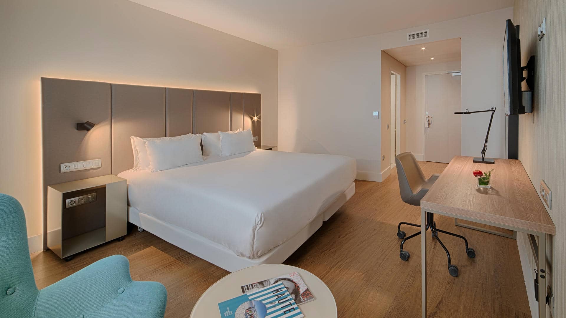 Chambre d’hôtel spacieuse avec un grand lit double aux draps blancs, une tête de lit capitonnée gris taupe et des appliques lumineuses intégrées. Au pied du lit, une table basse ronde blanche présente quelques magazines. À droite, un long bureau en bois clair est équipé d’une chaise de bureau grise, d’une lampe noire et d’un petit bouquet de fleurs rouges. Un téléviseur noir est fixé au mur au-dessus du bureau. À gauche, un fauteuil bleu turquoise est partiellement visible. Le sol est en parquet bois clair et les murs sont beige clair. Au fond, une porte donne sur l’entrée de la chambre.