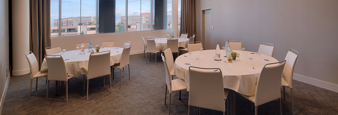 Salle de réunion aménagée pour un événement professionnel, comprenant plusieurs tables rondes recouvertes de nappes blanches et entourées de chaises rembourrées beige. Les tables sont équipées de bouteilles d’eau, de verres, de blocs-notes, de stylos et de petits arrangements floraux. De grandes fenêtres le long d’un mur apportent de la lumière naturelle et offrent une vue sur des bâtiments modernes à l’extérieur. Une moquette gris foncé recouvre le sol, tandis que les murs clairs et le mobilier aux tons neutres créent une atmosphère professionnelle et sobre. Des rideaux marron encadrent les fenêtres et complètent les tons chaleureux de l’intérieur. Une porte est visible au fond de la salle.